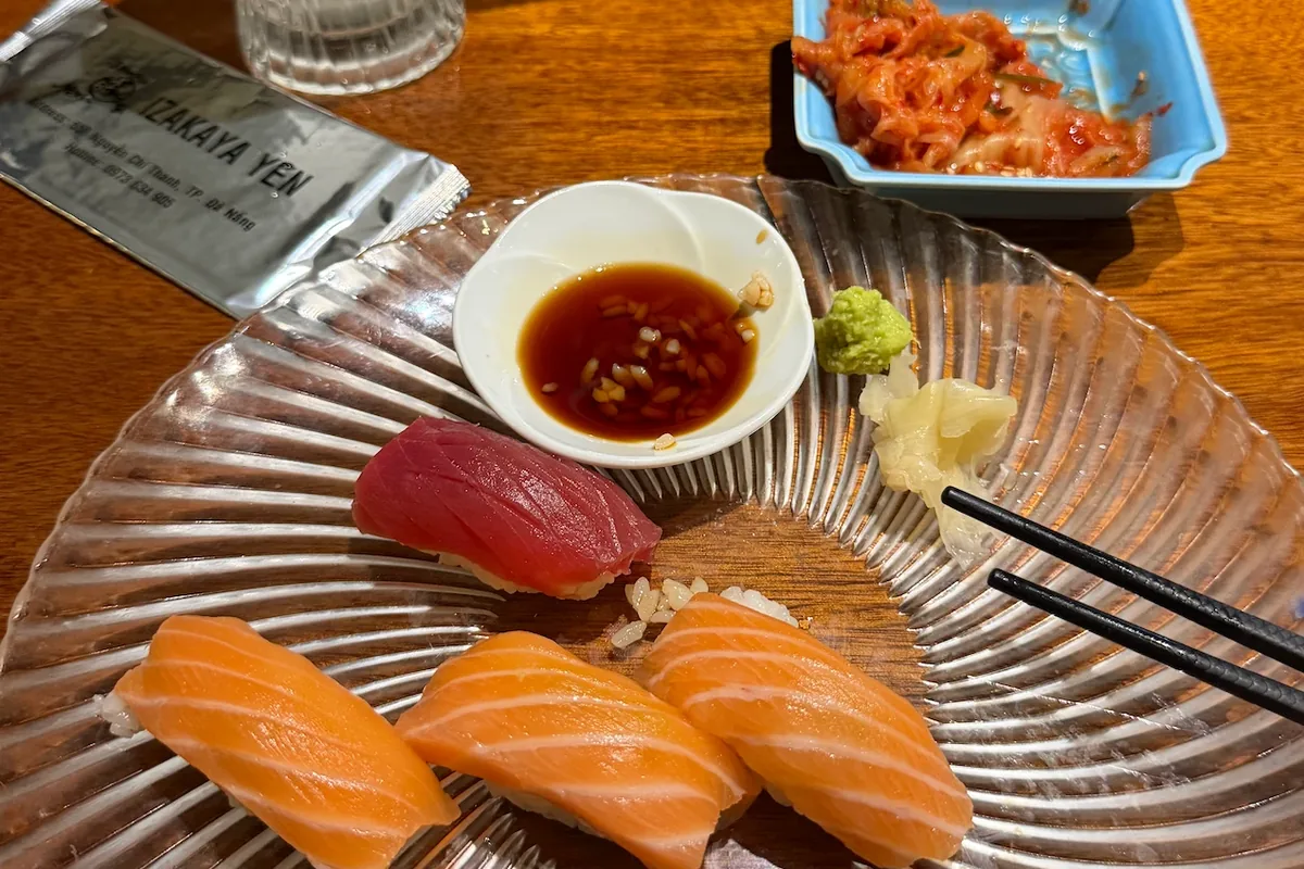 Three pieces of salmon nigiri and one of tuna on a ribbed glass plate, with soy sauce, wasabi, pickled ginger and a dish of kimchi