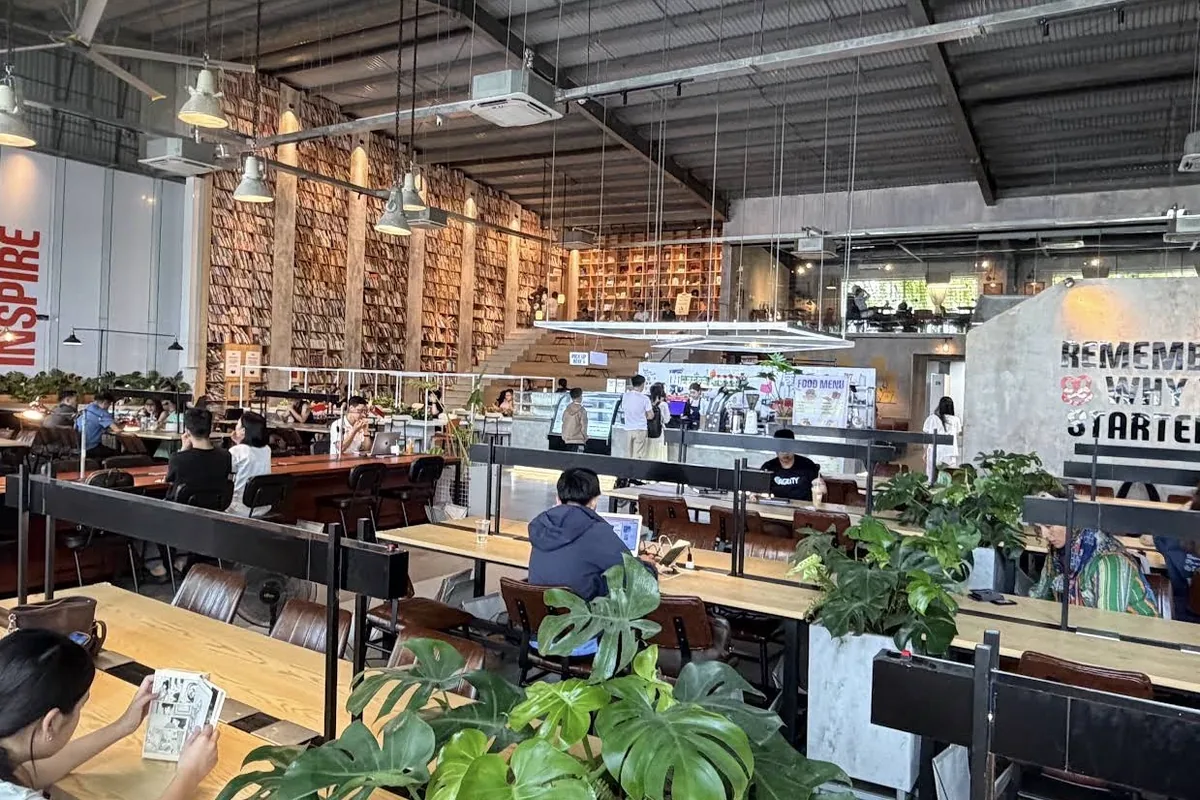 A high-ceilinged industrial café with long shared tables, a wall of shelved books and plants between the desks