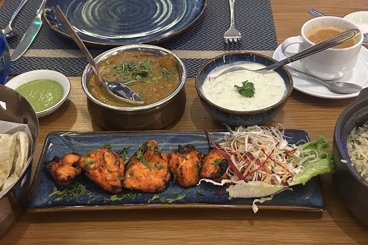 Tandoori chicken on a long blue platter with a bowl of curry, raita, naan, rice and a cup of chai