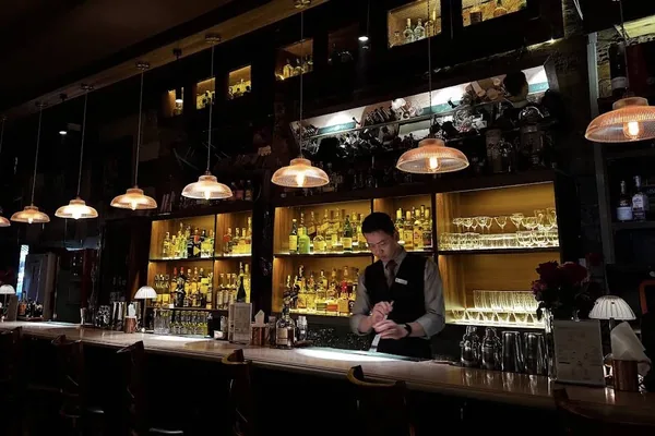 A bartender at work behind the long bar under a row of pendant lamps, with backlit shelves of spirits