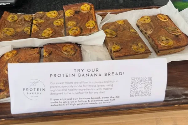Slices of protein banana bread topped with baked banana rounds on a wooden board, beside the bakery’s flyer