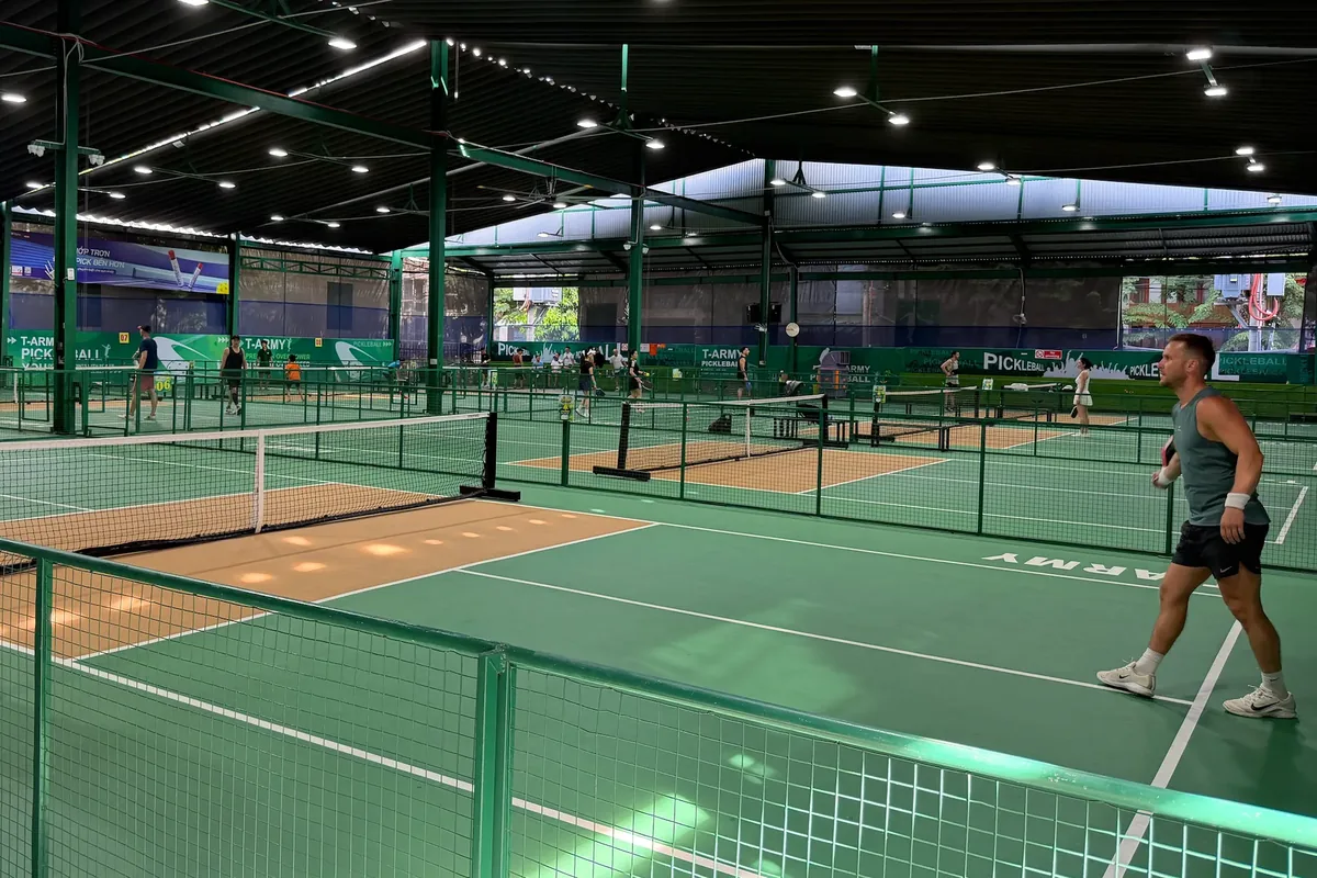 Covered pickleball courts with green fencing and games in play, and a player walking to the baseline