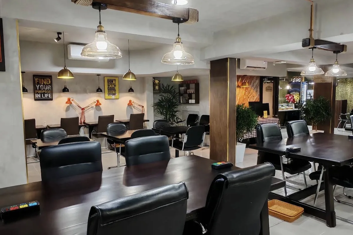 Long dark wooden tables with black leather chairs under glass pendant lamps hung from a rope-bound beam, with orange desk lamps and framed posters on the back wall
