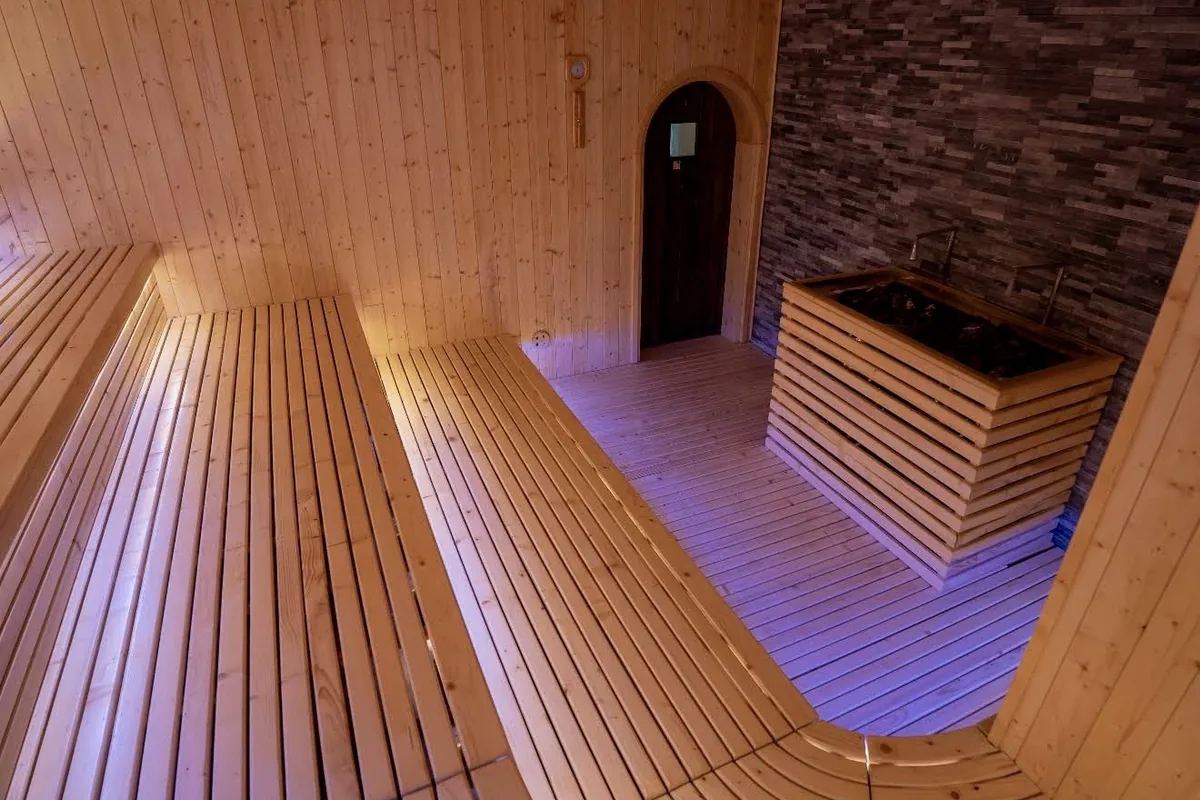 Inside a pale timber sauna: two tiers of slatted benches, a slatted box of heater stones against a grey stone wall, and an arched dark door
