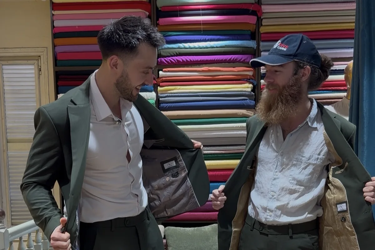 Jake and a friend showing the linings of their new green suits in front of a wall of fabric bolts